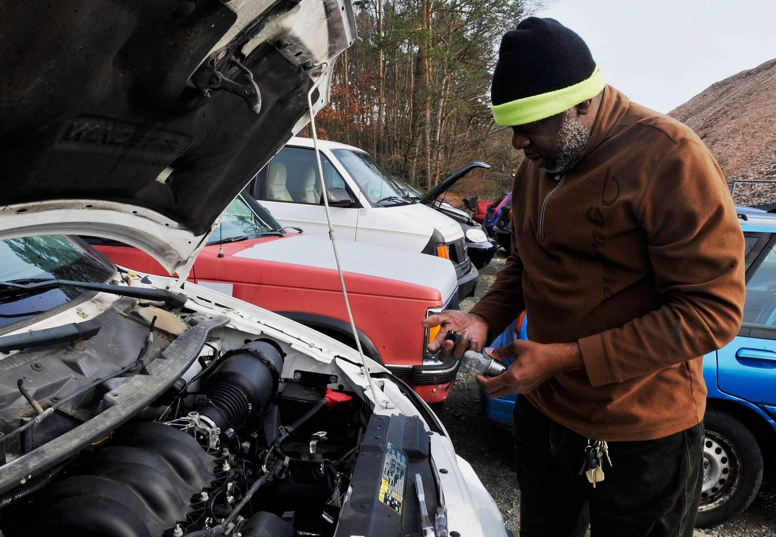 Black man wearing a brown jacket and knit cap removes a part from an automobile engine