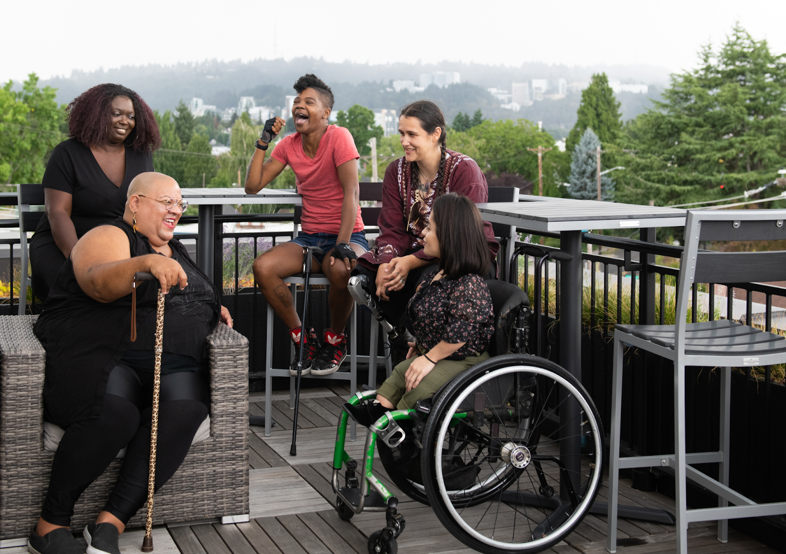 Five people of color with canes, prosthetic legs, and a wheelchair sitting on a rooftop deck, talking