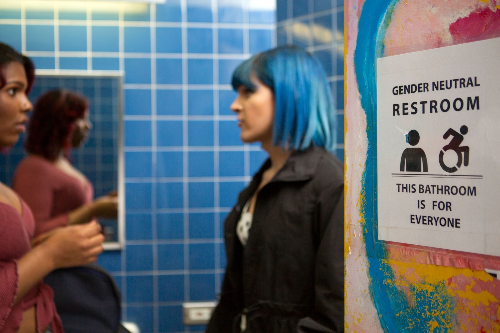 Two people who appear feminine in conversation near a sign: "gender-neutral bathroom. This bathroom is for everyone."