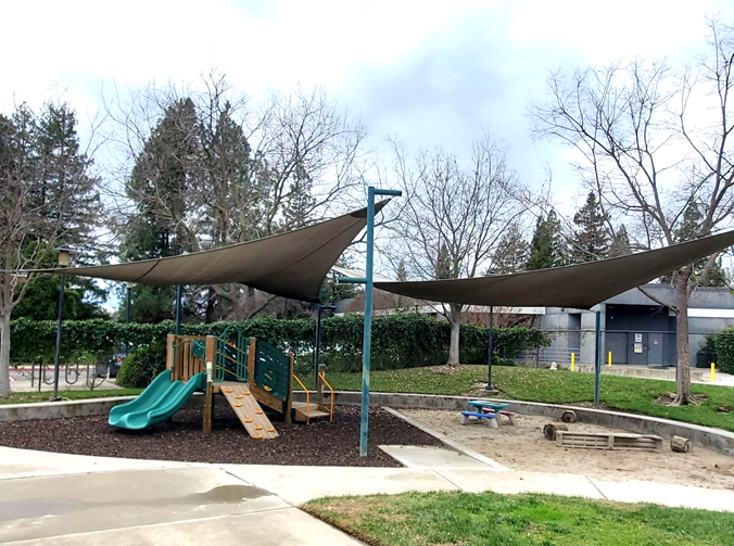 Outdoor play yard for toddlers with large climbing structure and low picnic table and shade structures overall all and encircled with cement. 