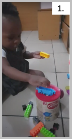 Nora has a yellow block in her left hand and a blue block in her right hand, but is primarily focused on the blue block. In front of her is the cylinder container with a hole in its lid. and various other blocks scattered around the base of the cylinder. With the blue block in her right hand, Nora moves it toward the hole in the cylinder container and drops it onto the container lid. When laid horizontally on top of the container, the blue block is too long to fit through the round hole.