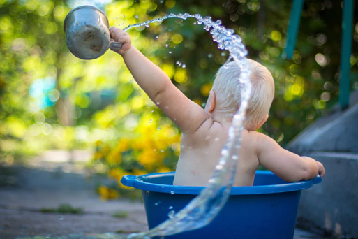 Infant sitting in small plastic tub pouring water from a metal bucket outside of in a pool of water
