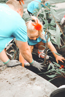 Caregiver and toddler side by side in a garden. Toddler is moving dirt with hands onto newly planted tree while caregiver assists. 