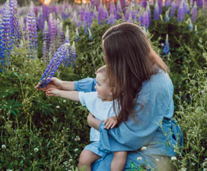 Caregiver holds older infant up and both grasp large lilac flower. 