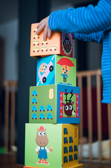 Toddler stands and uses both hands to add fourth block to stack of blocks