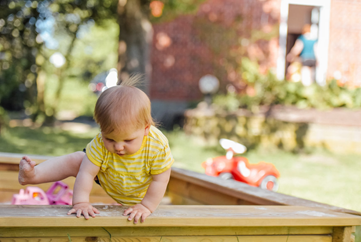 Child attempting to climb out of a sandbox by lifting left leg over ledge and grasping with both hands in front of body. 