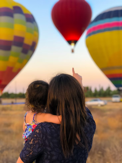 Caregiver holding toddler in arms pointing to hot air balloon as toddler looks on.
