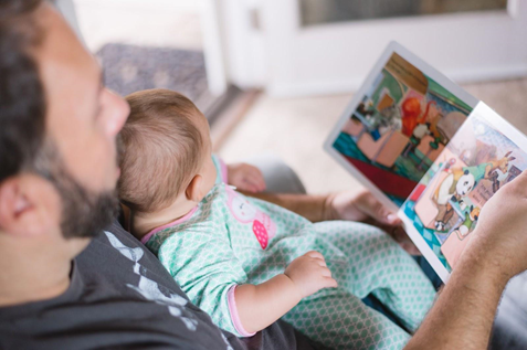 Caregiver holding infant in lap with open book, reading to infant. 
