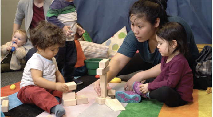 Caregiver sitting between two seated toddlers all stacking blocks. 