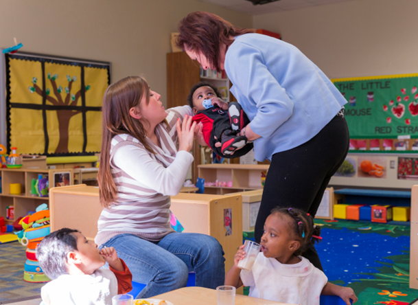 Infants and toddlers in a mixed-age classroom. Caregivers handing off young infant with two toddlers in foreground at table. 