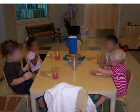 Niños pequeños sentados en la mesa baja comiendo merienda. Las tazas y los platos se colocan antes de cada uno con una jarra en el medio.