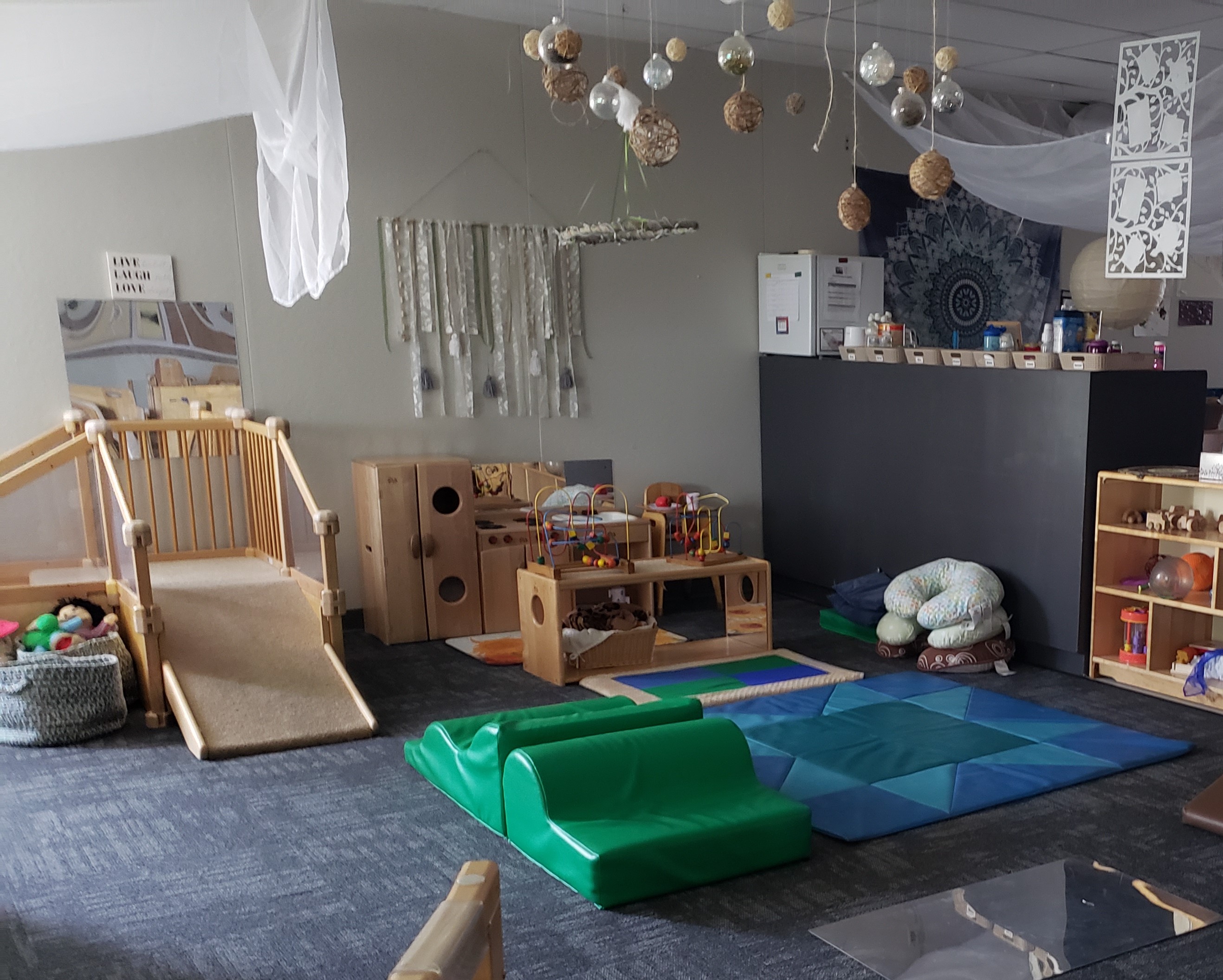 Ambiente infantil que contiene una pequeña estructura de escalada, cocina de juegos de madera, esteras de plástico en el piso y una pequeña estructura de escalada de espuma.