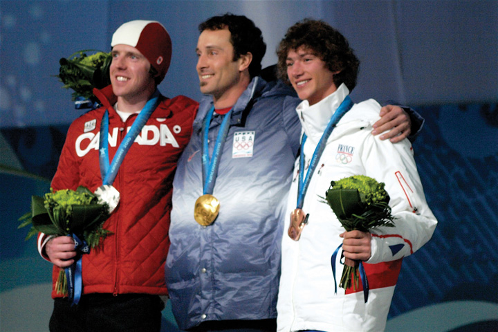 Three male, smiling Olympic athletes posing for photos holding flowers with a gold, silver, and bronze medal around their necks.