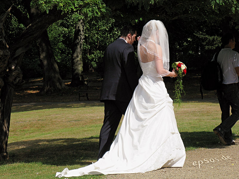 couple in a park with young man in a suit and young woman in a long wedding dress and holding wedding bouquet.