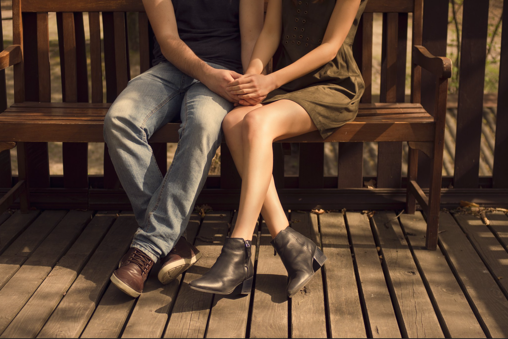 Joven pareja romántica sentada cerca en un banco al aire libre, tomados de la mano íntimamente; hombre vistiendo jeans, mujer una falda corta.