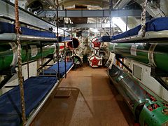 Photo shows inside a submarine torpedo room, with sailors' bunks on one wall, torpedoes on the other, tubes straight ahead.