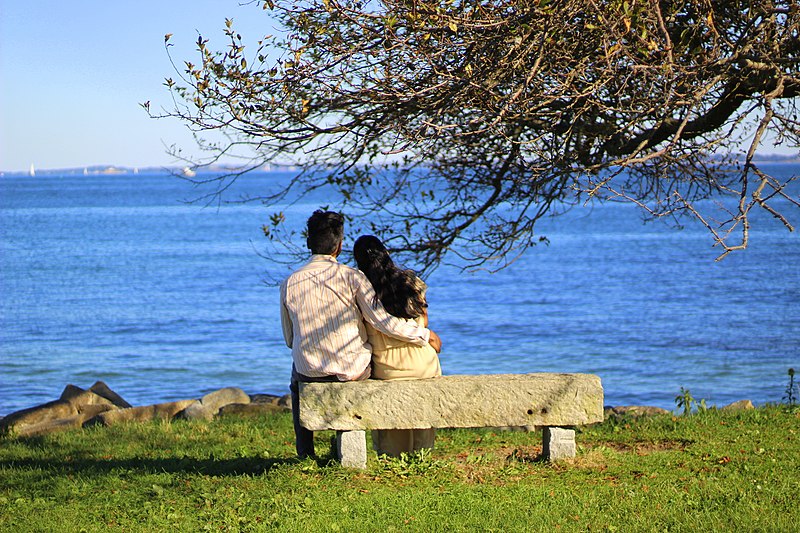 Photo behind a young couple sitting close on a stone bench facing the sea; young man's arm around woman leaning to him.