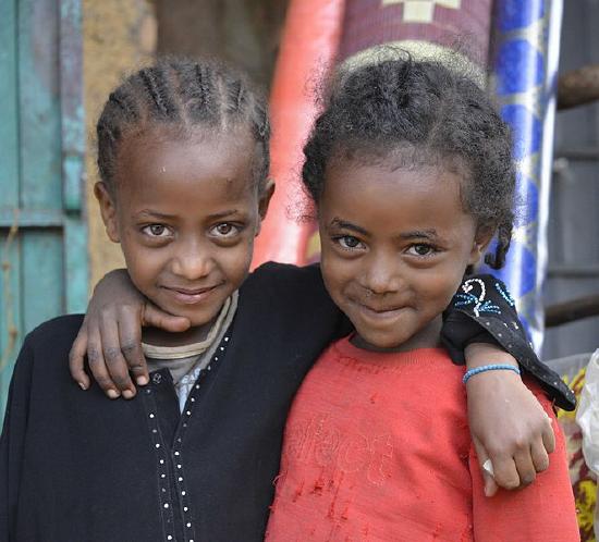 Photo of young friends; two cute black girls, 7 or 8 years old, each smiling and with one arm over the shoulder of the other.