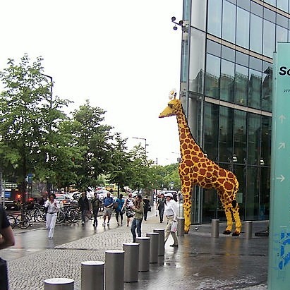 Un modelo de tamaño real de una jirafa se encuentra en una concurrida plaza pública.