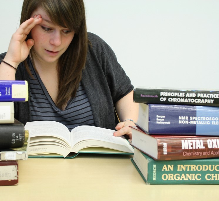 Una estudiante lee un libro de texto grande con pilas de otros libros de texto sobre la mesa a su alrededor.