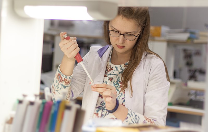 Young woman chemist in white lab coat, surrounded by laboratory equipment, is filling a test tube with liquid using a pipette.