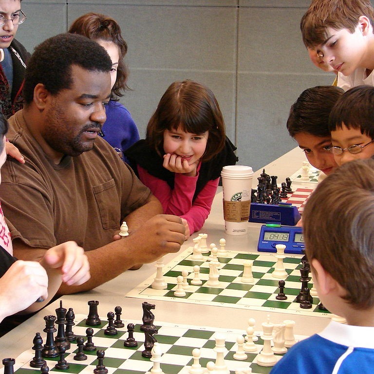 Foto que muestra a un grupo de niños de pie, observando atentamente a un adulto jugando una partida de ajedrez.