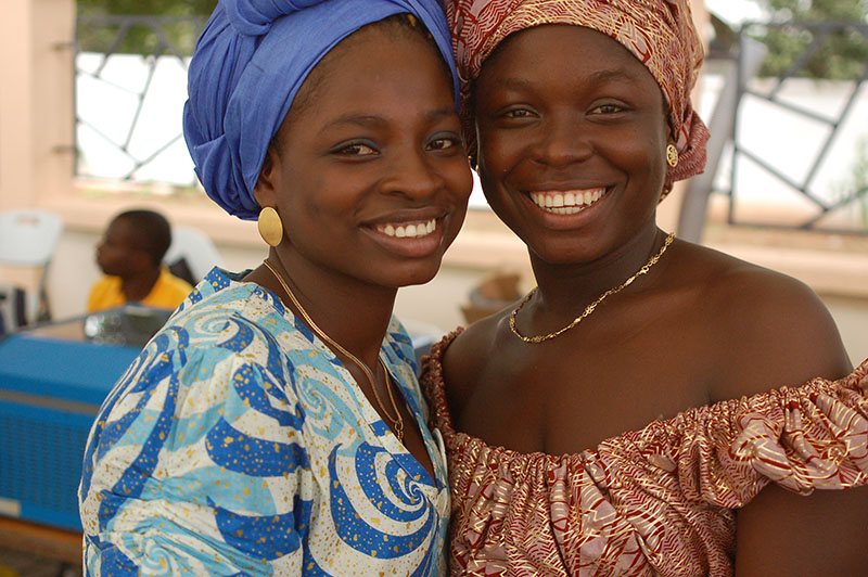 两名加纳妇女站着，头朝前触摸。 两人都在微笑。 左边的女人戴着一条蓝色的头巾和一件蓝色图案的连衣裙。 右边的女人戴着一条橙色图案的头巾和一件相同图案的露肩连衣裙。 两位女士都戴着简单的金链项链和耳环。