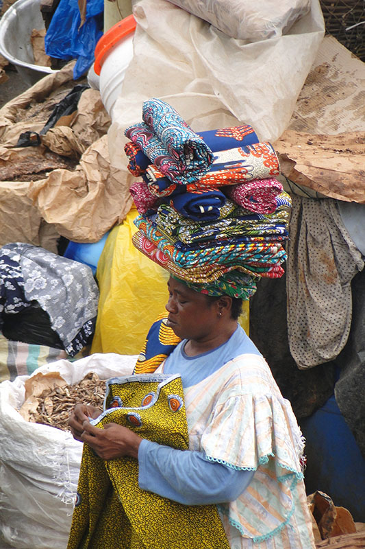 Une Ghanéenne debout sur un étal de marché remplie de balles de vêtements. Elle porte une robe blanche et bleue et a une pile de tissu plié coloré sur la tête. Elle plie un morceau de tissu jaune.